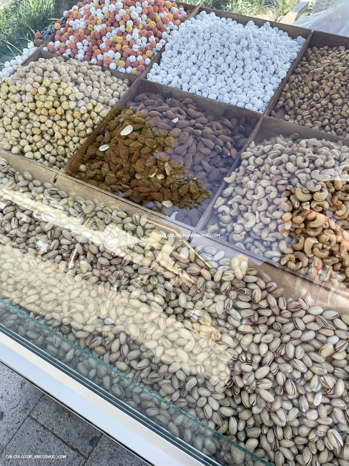 Витрина с различными видами орехов: фисташки, кешью, миндаль и нут в сахаре, аккуратно разложенные по деревянным ячейкам.