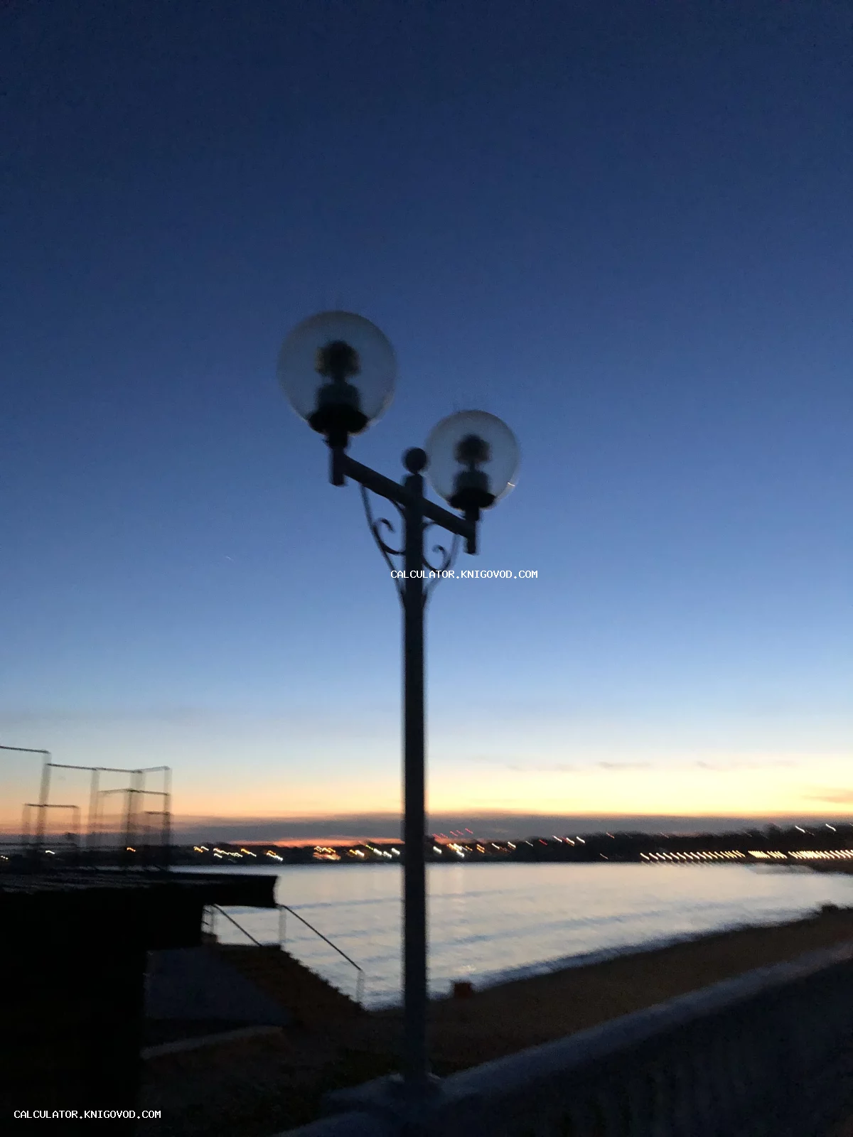Уличный фонарь с двумя шарообразными плафонами на фоне вечернего неба и морского берега в час заката.