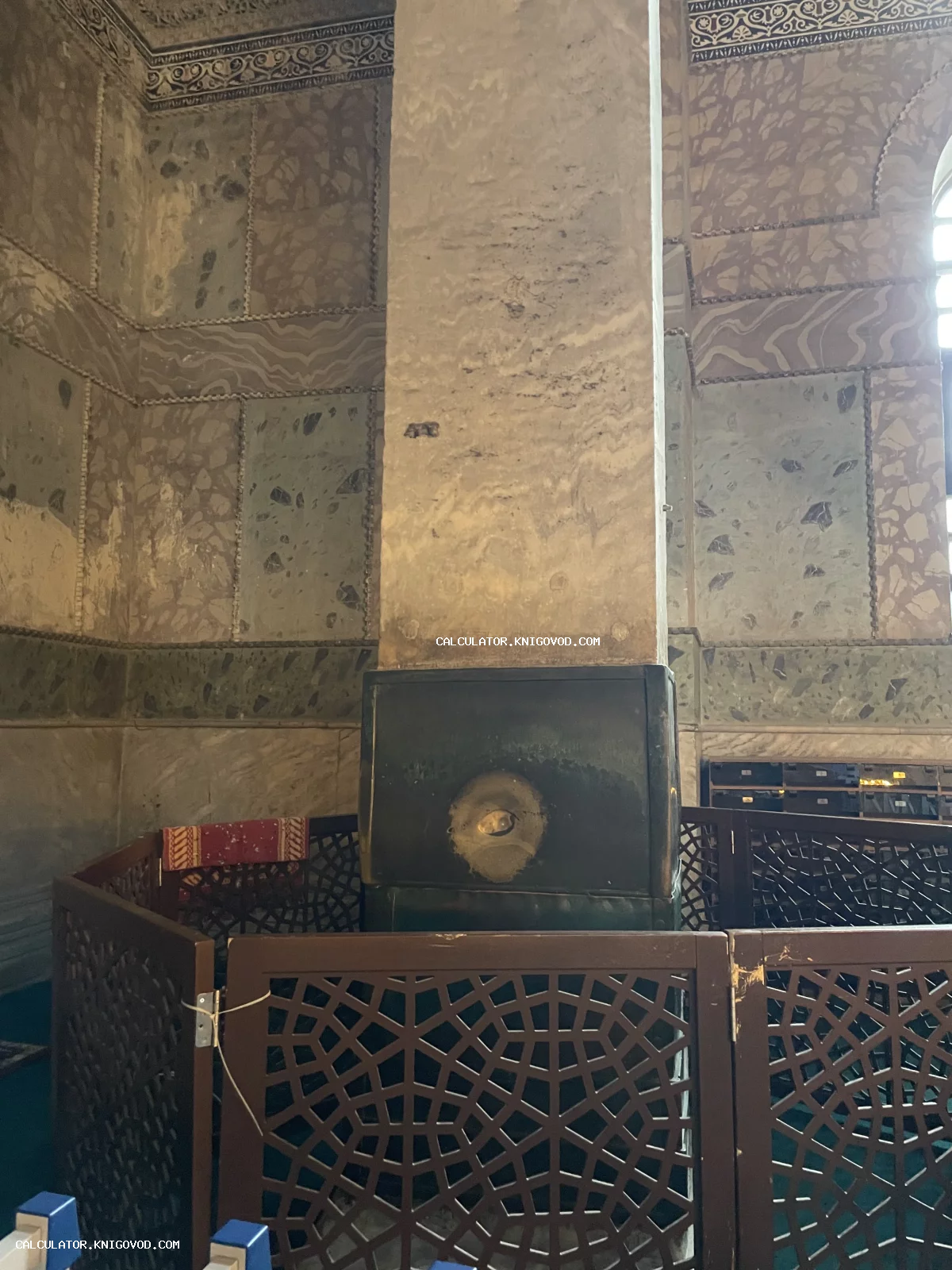 Мраморная колонна с медной облицовкой и отверстием в центре, окруженная деревянной резной оградой в интерьере мечети Айя-София.