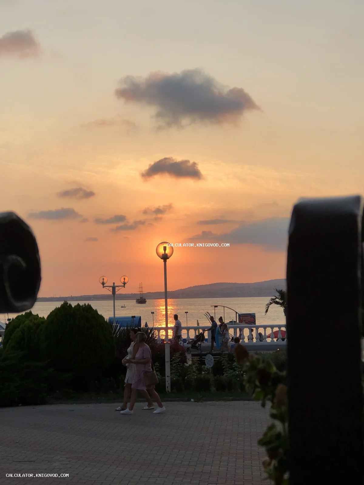 Вид на морской закат через кованую ограду, на переднем плане гуляющие по набережной люди, уличные фонари и белая балюстрада на фоне оранжевого неба.