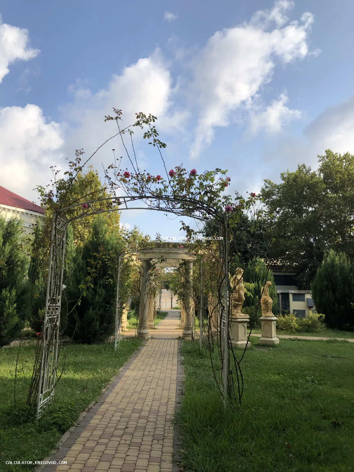 Садовая дорожка, проходящая через кованую цветочную арку и каменную колоннаду со статуями на фоне деревьев и облачного неба.