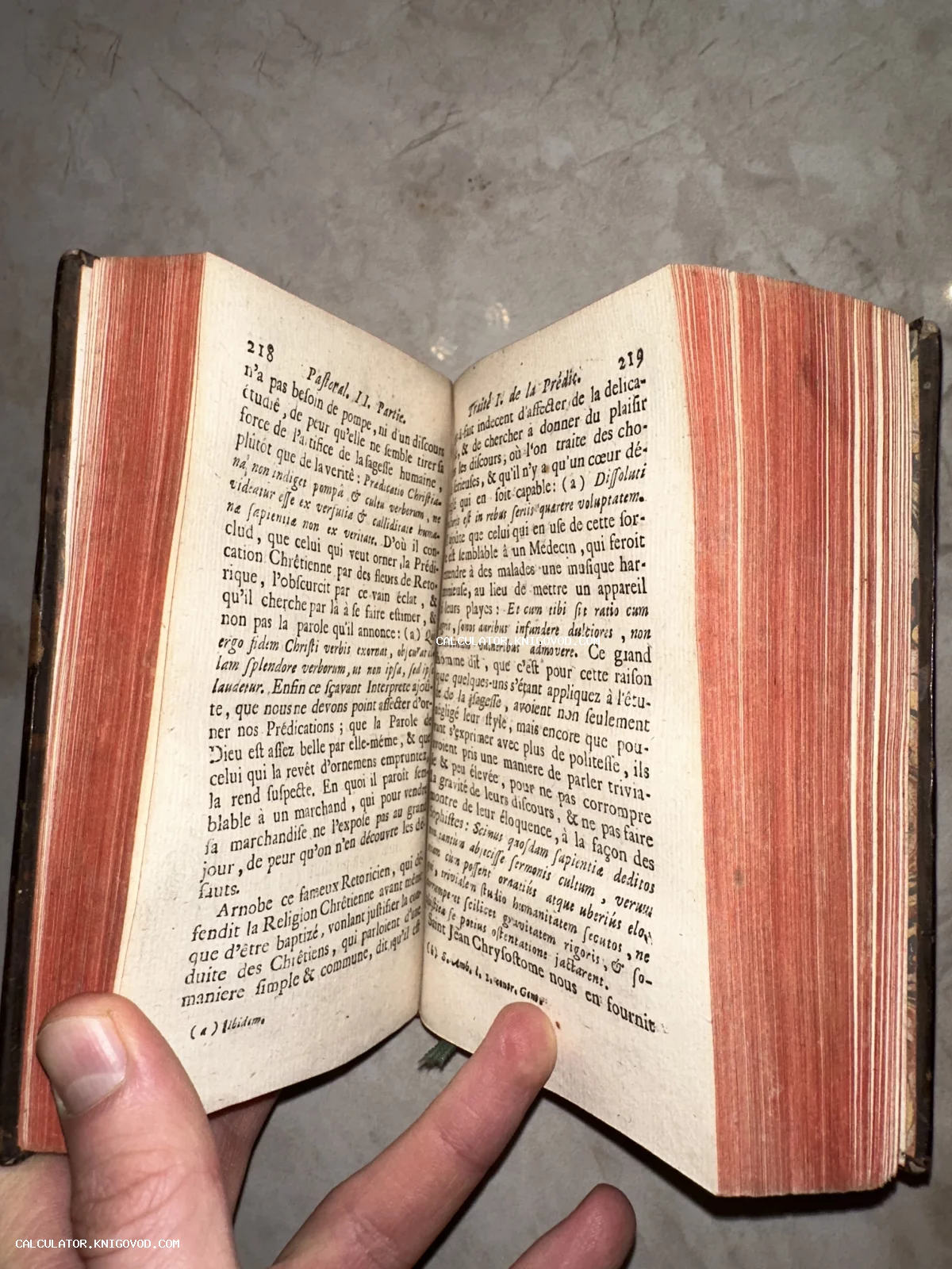 Раскрытая старинная книга на французском языке с пожелтевшими страницами и красным тонированным обрезом, удерживаемая рукой.