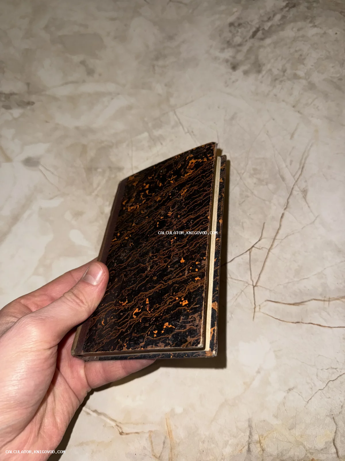 Старинная карманная книга с темной обложкой, украшенной оранжевым мраморным узором, в руке человека на фоне светлого мрамора.