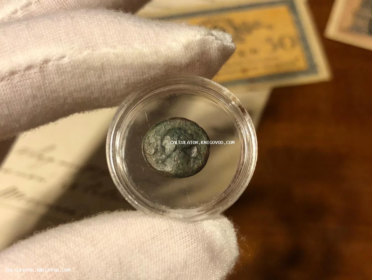 Рука в белой перчатке держит маленькую античную монету с патиной в защитной пластиковой капсуле на фоне старых документов.