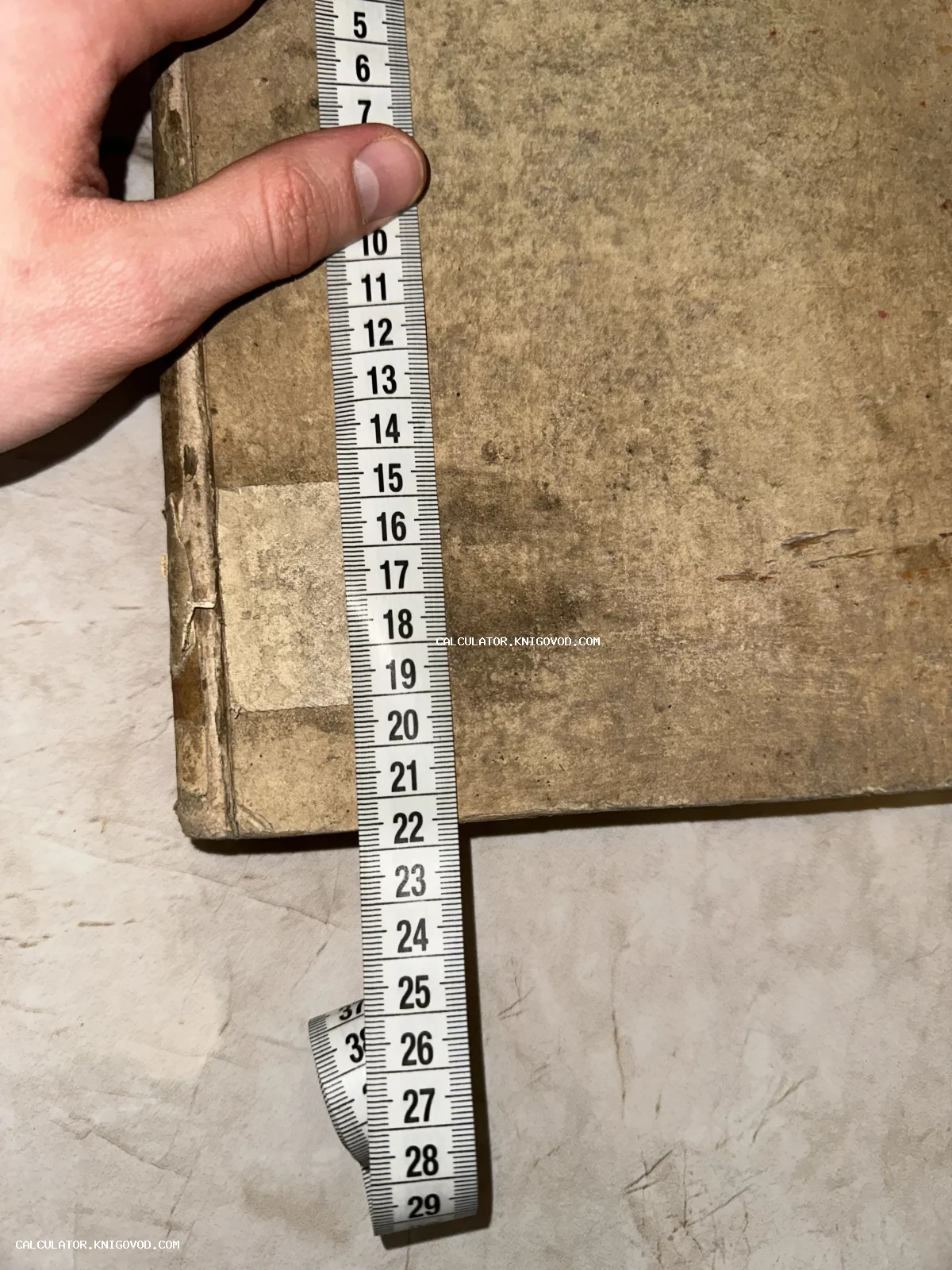 Процесс измерения высоты старинной книги в потертом картонном переплете с помощью сантиметровой ленты.