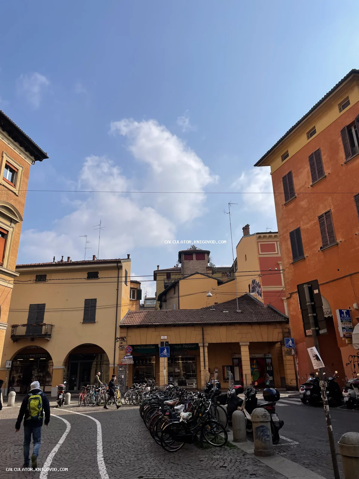 Вид на небольшую городскую площадь в Италии с историческими зданиями охристого цвета, парковкой для велосипедов и прохожими под ясным небом.