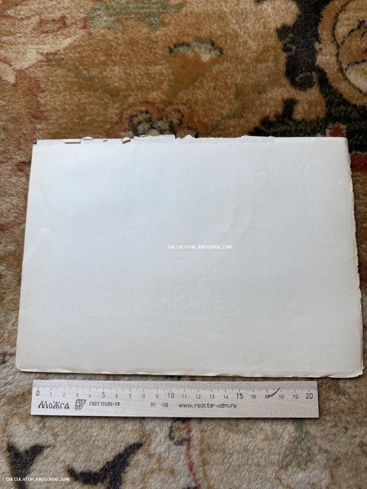 Пустой пожелтевший лист старой бумаги или картона с неровными краями на фоне ковра, внизу лежит деревянная линейка.