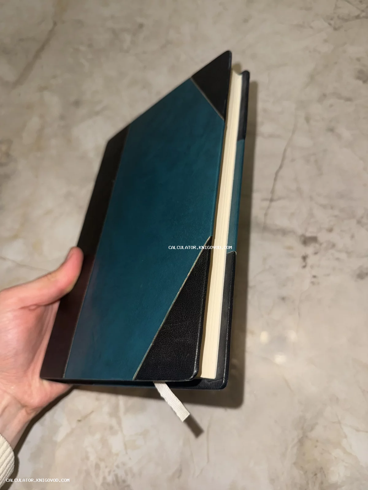 Рука держит закрытую книгу в бирюзово-черной обложке с закладкой на фоне мраморной поверхности.
