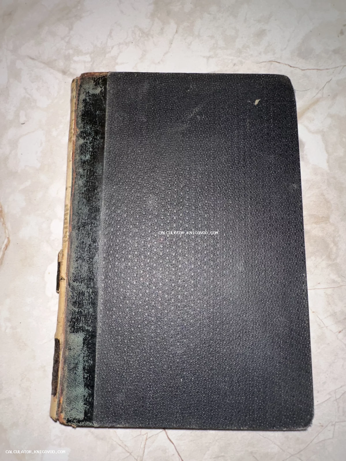 Старинная книга с темной текстурированной обложкой и поврежденным корешком на светлом мраморном фоне.