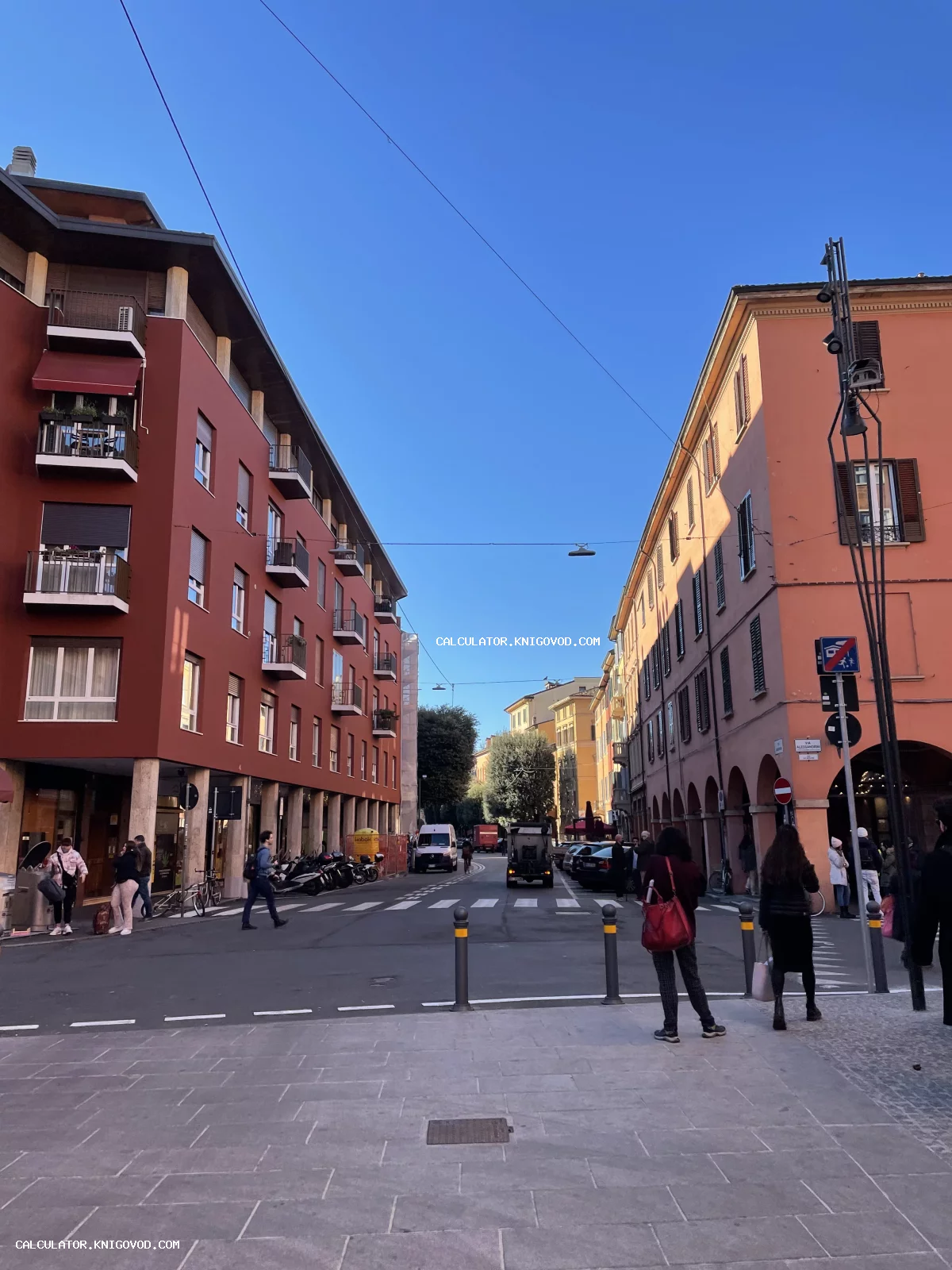 Вид на городскую улицу в Болонье с характерными терракотовыми зданиями, аркадами и прохожими под ясным синим небом.