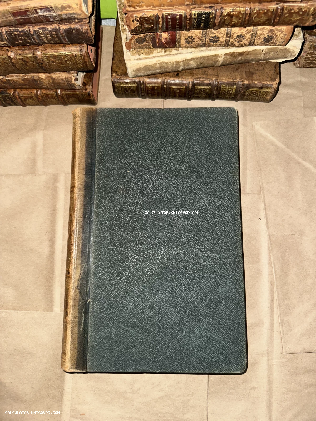 Антикварная книга с темно-зеленой фактурной обложкой на фоне стопок старинных книг с золотым тиснением на корешках.