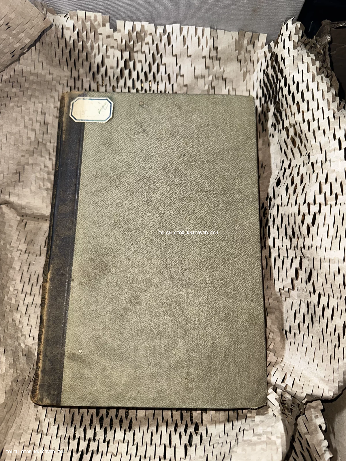 Старинная книга с серой текстурированной обложкой и темным корешком, лежащая на защитной упаковочной бумаге.