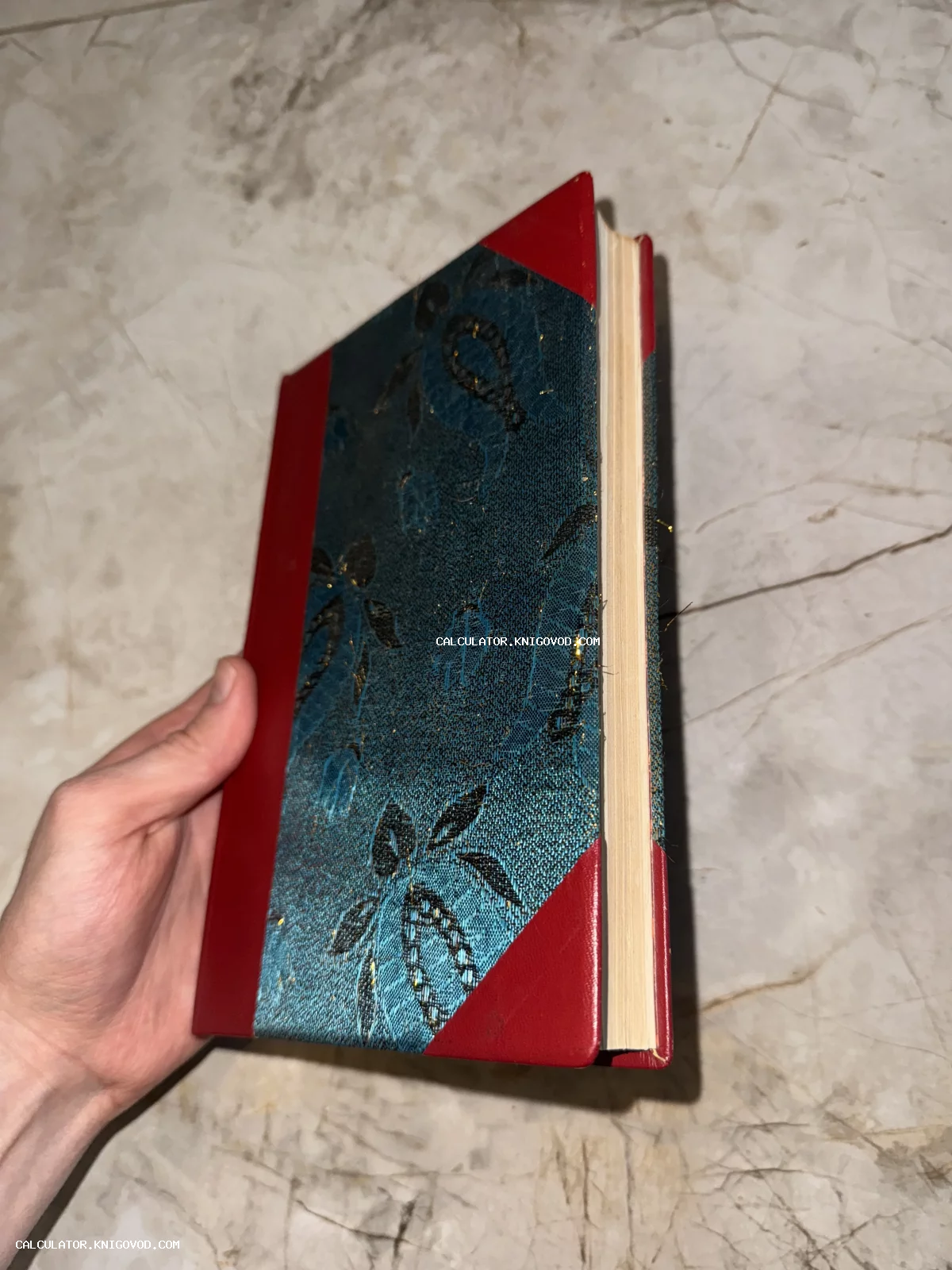 Книга в руке с комбинированным переплетом из красной кожи и бирюзовой узорчатой ткани с золотыми нитями.