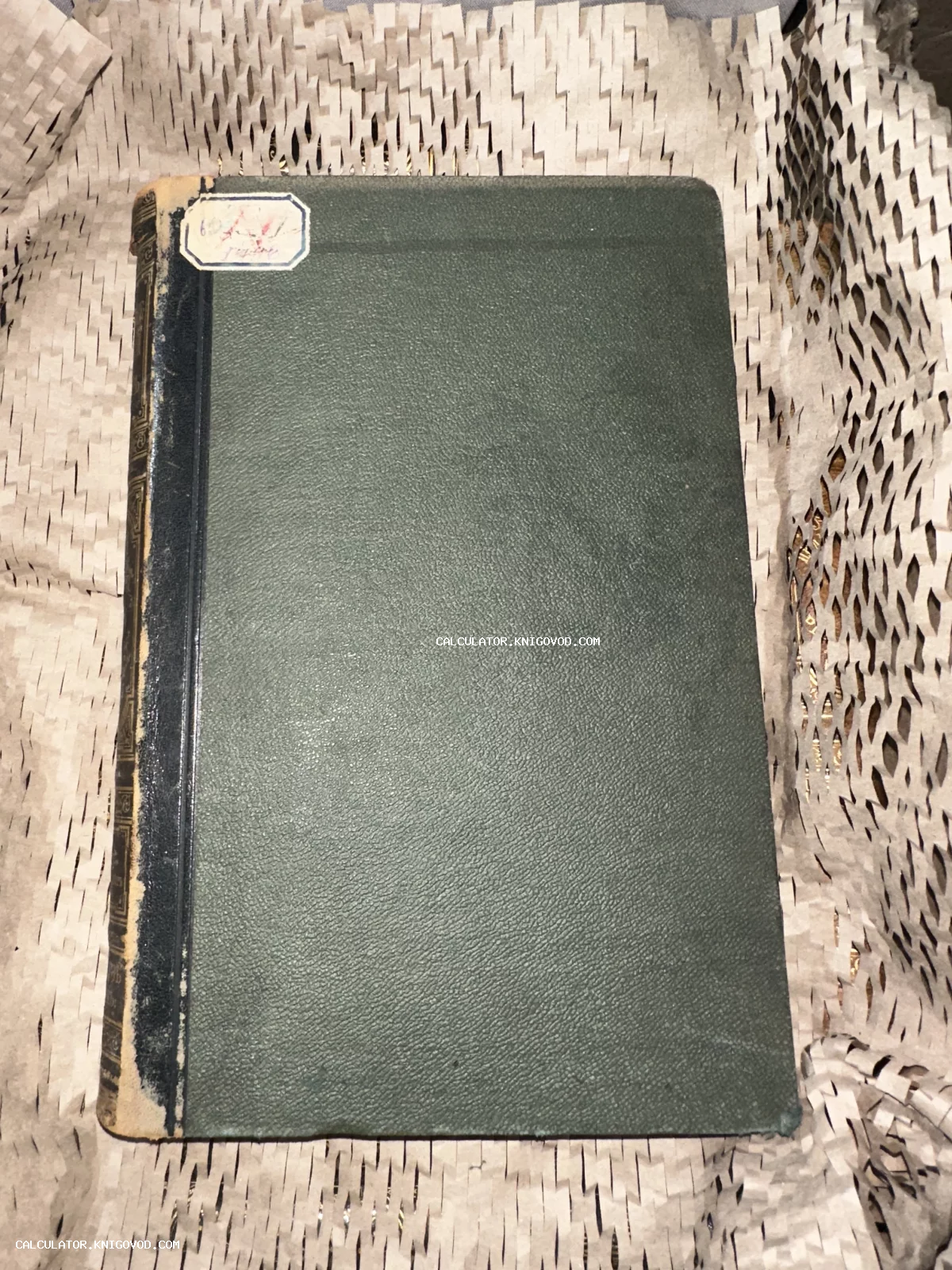 Вид сверху на антикварную книгу с темно-зеленой фактурной обложкой, потертым темным корешком и бумажным ярлыком в углу.