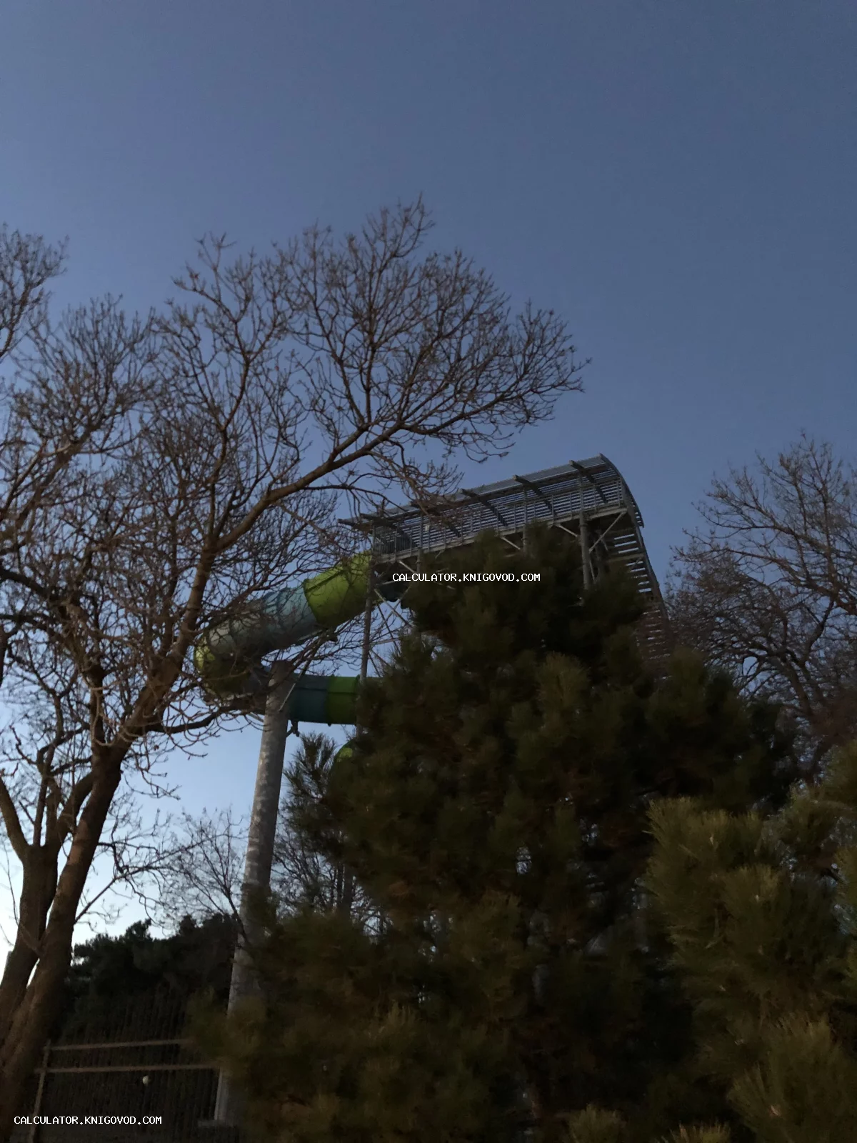 Конструкция водной горки и деревянная смотровая площадка среди деревьев на фоне темно-синего вечернего неба.