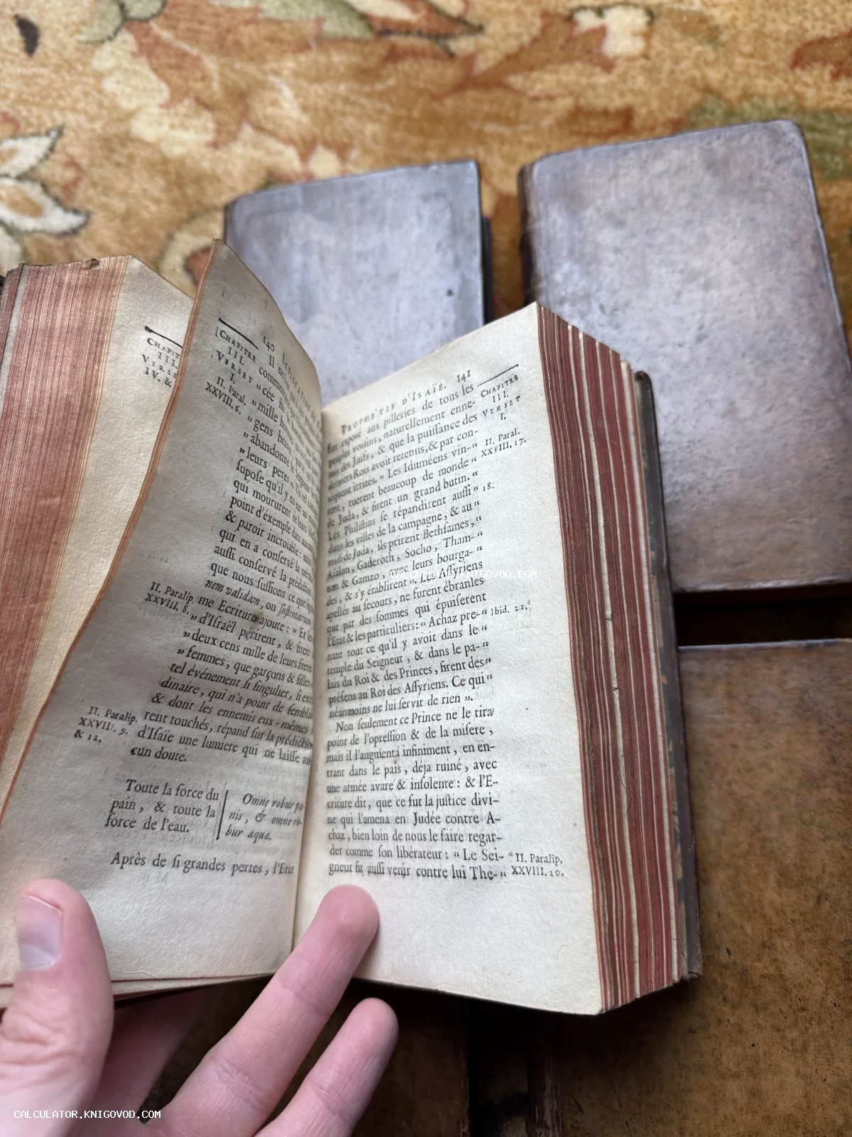 Раскрытая антикварная книга на французском языке с красным тонированным обрезом на фоне других старинных томов.