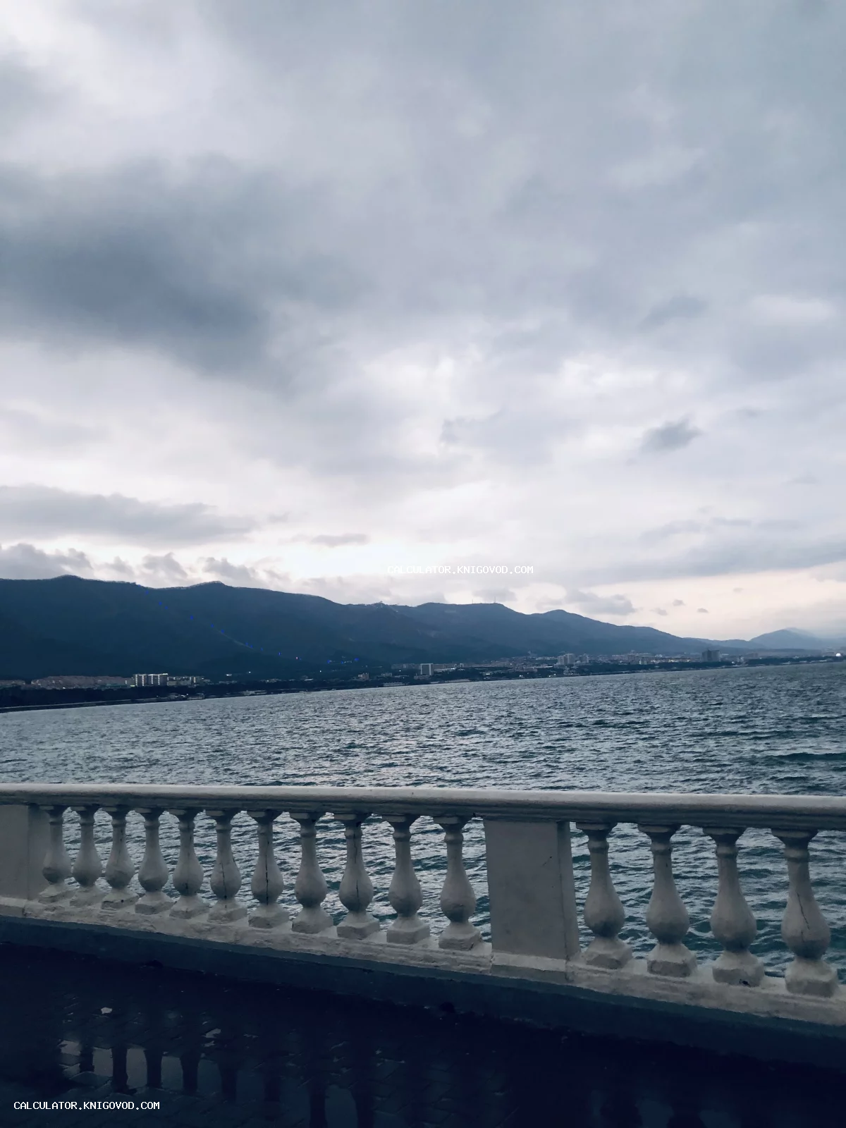 Вид на море и горы с набережной через белую каменную балюстраду под пасмурным небом.