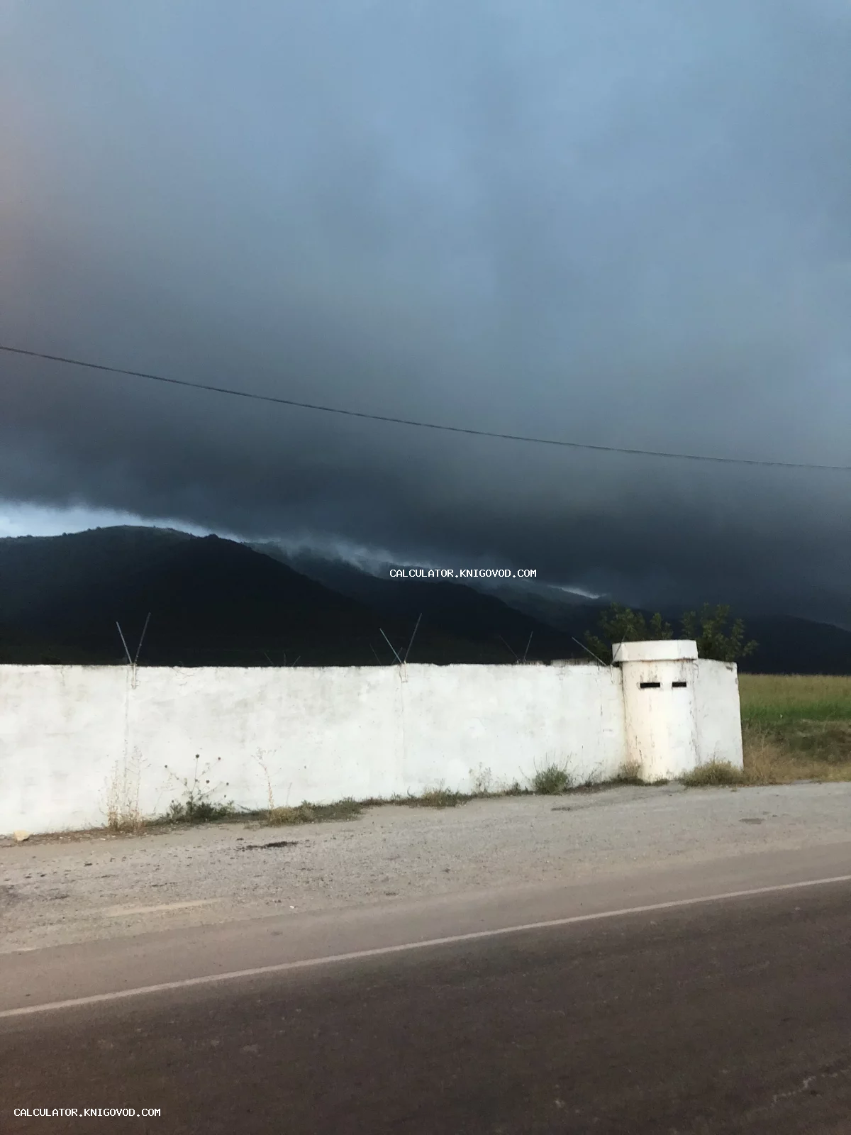 Белая бетонная стена с дозорной вышкой на фоне темных грозовых облаков и силуэтов гор.