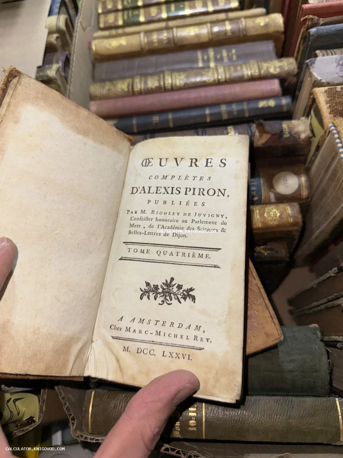 Разворот антикварной книги 'Oeuvres Completes d'Alexis Piron' 1776 года издания на фоне множества старинных томов в кожаных переплетах.