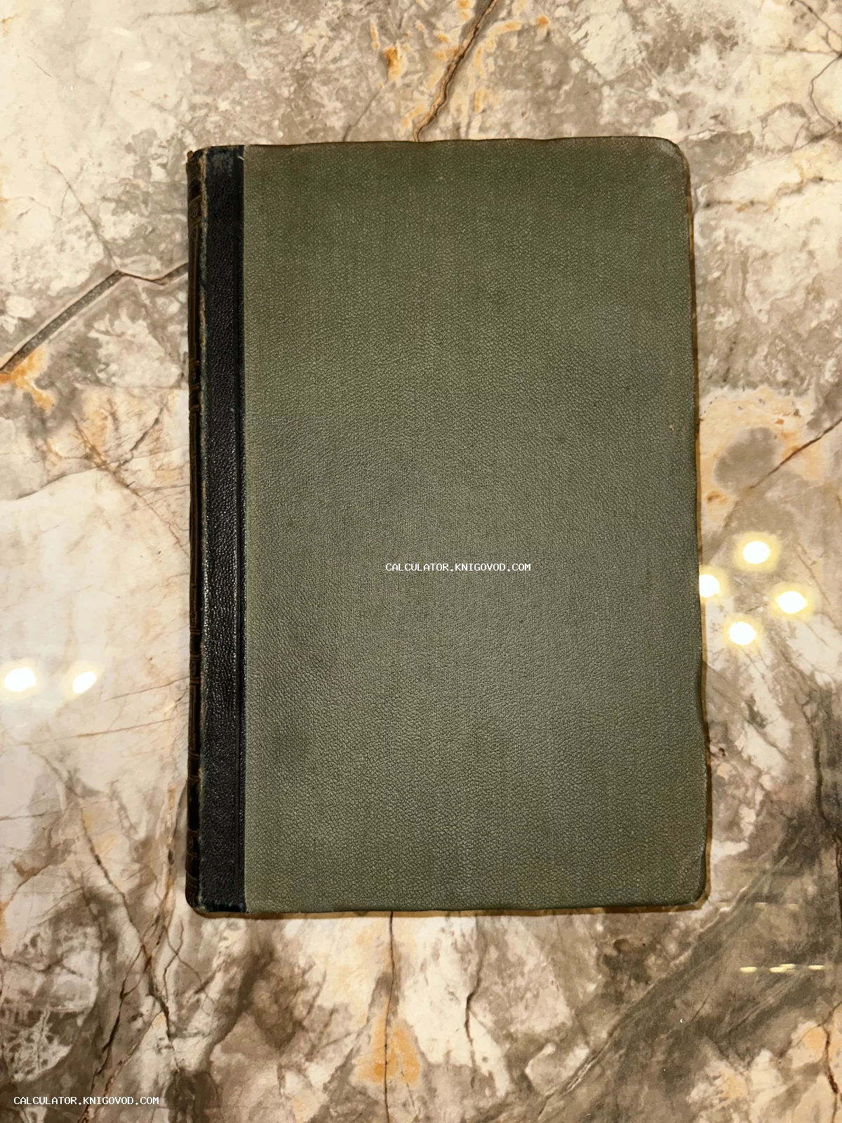Антикварная книга с темно-зеленой фактурной обложкой и черным корешком на мраморном столе.