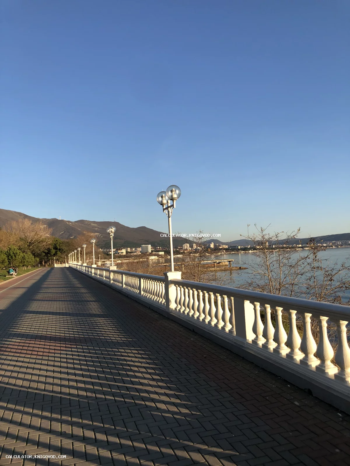 Пешеходная набережная с белой балюстрадой, уличными фонарями и видом на морской залив и горы под чистым небом.