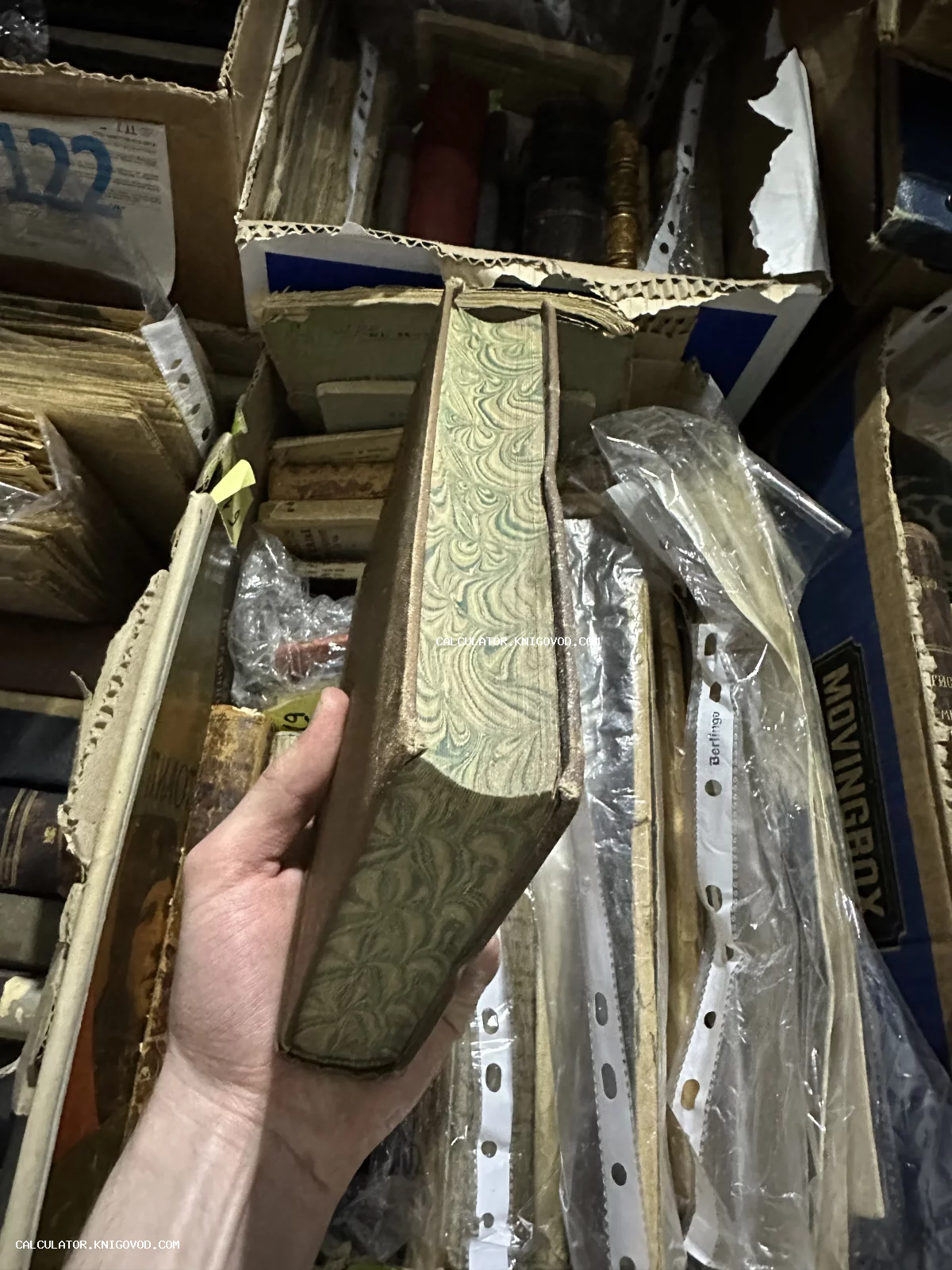 Старинная книга с декоративным мраморным обрезом в руке коллекционера на фоне архивных коробок с литературой.