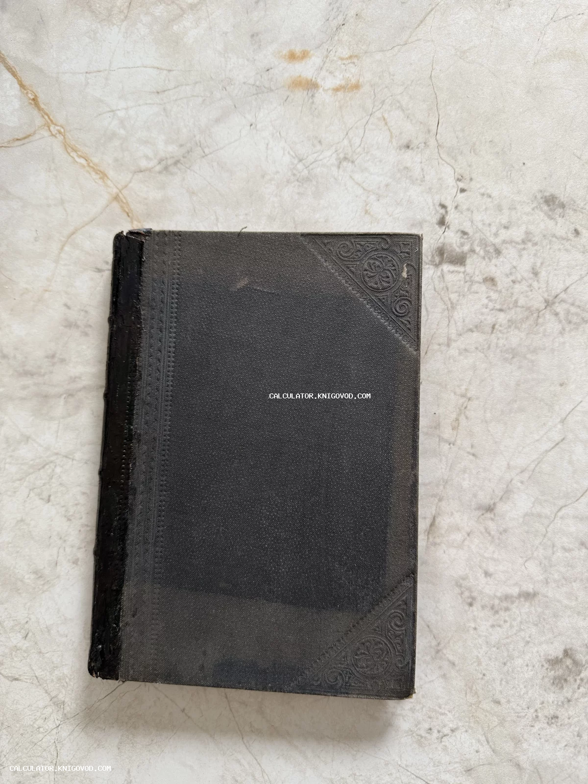 Старинная книга с темной фактурной обложкой и орнаментальным тиснением на углах, лежащая на светлой поверхности.