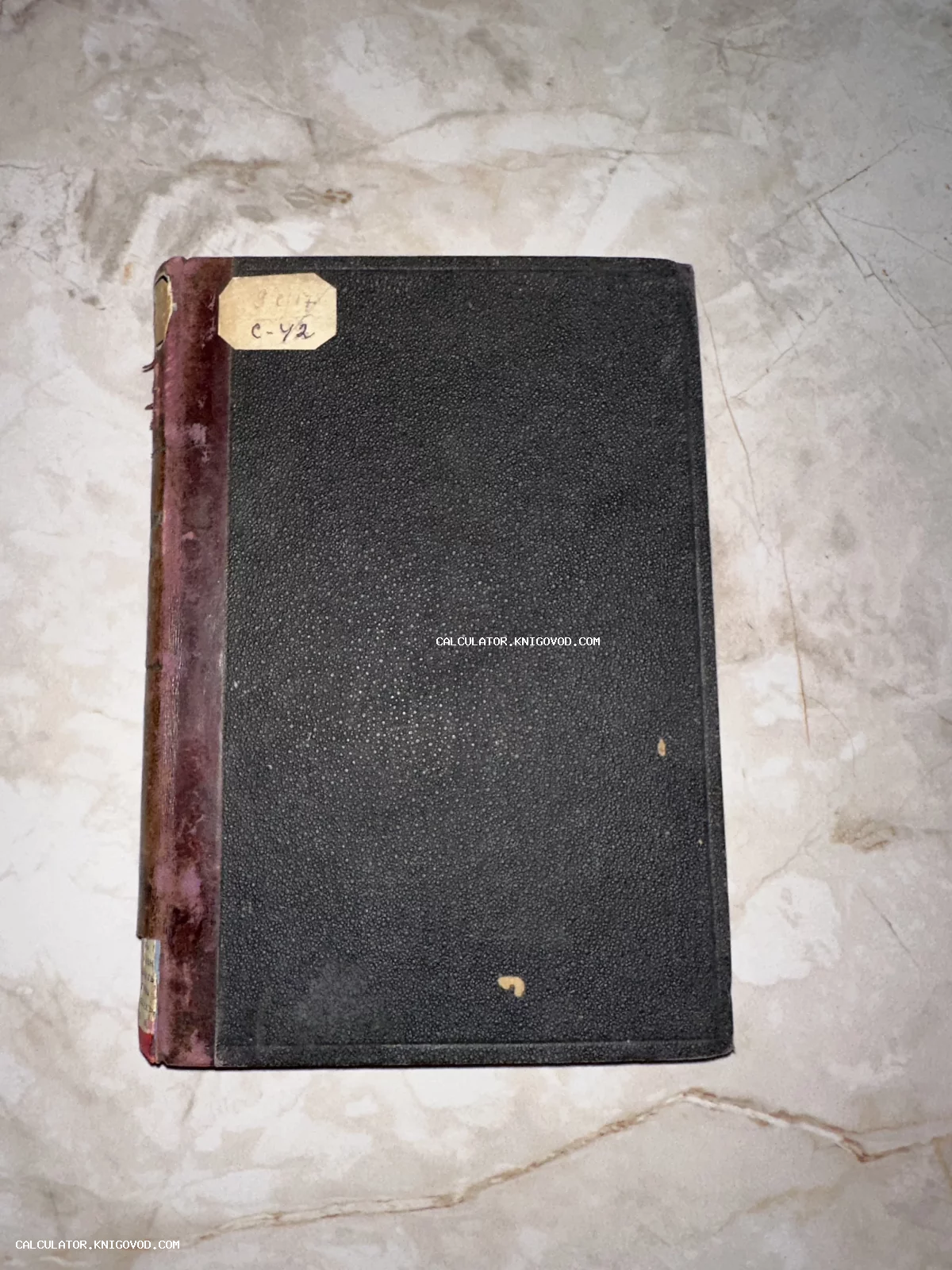 Старинная книга в темном фактурном переплете с потертым бордовым корешком на мраморной поверхности.