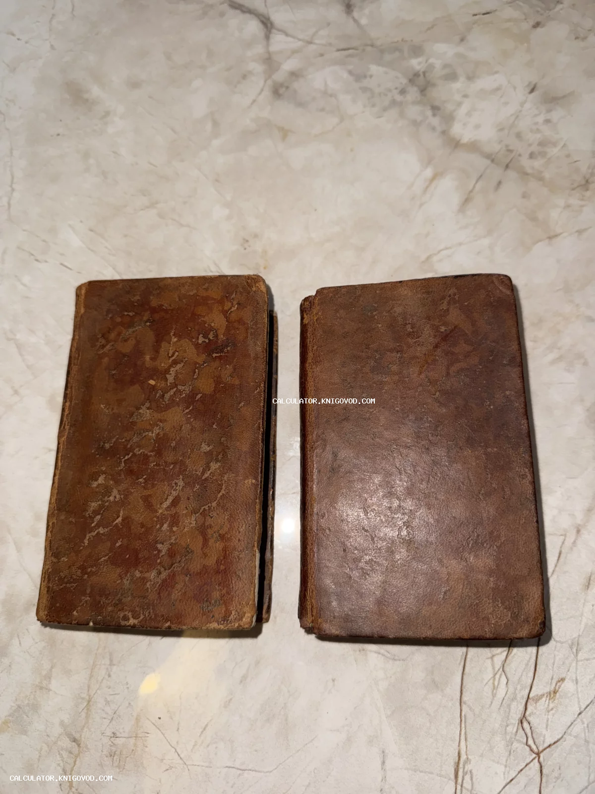 Две старинные малоформатные книги в потертых коричневых кожаных переплетах на светлой мраморной поверхности.