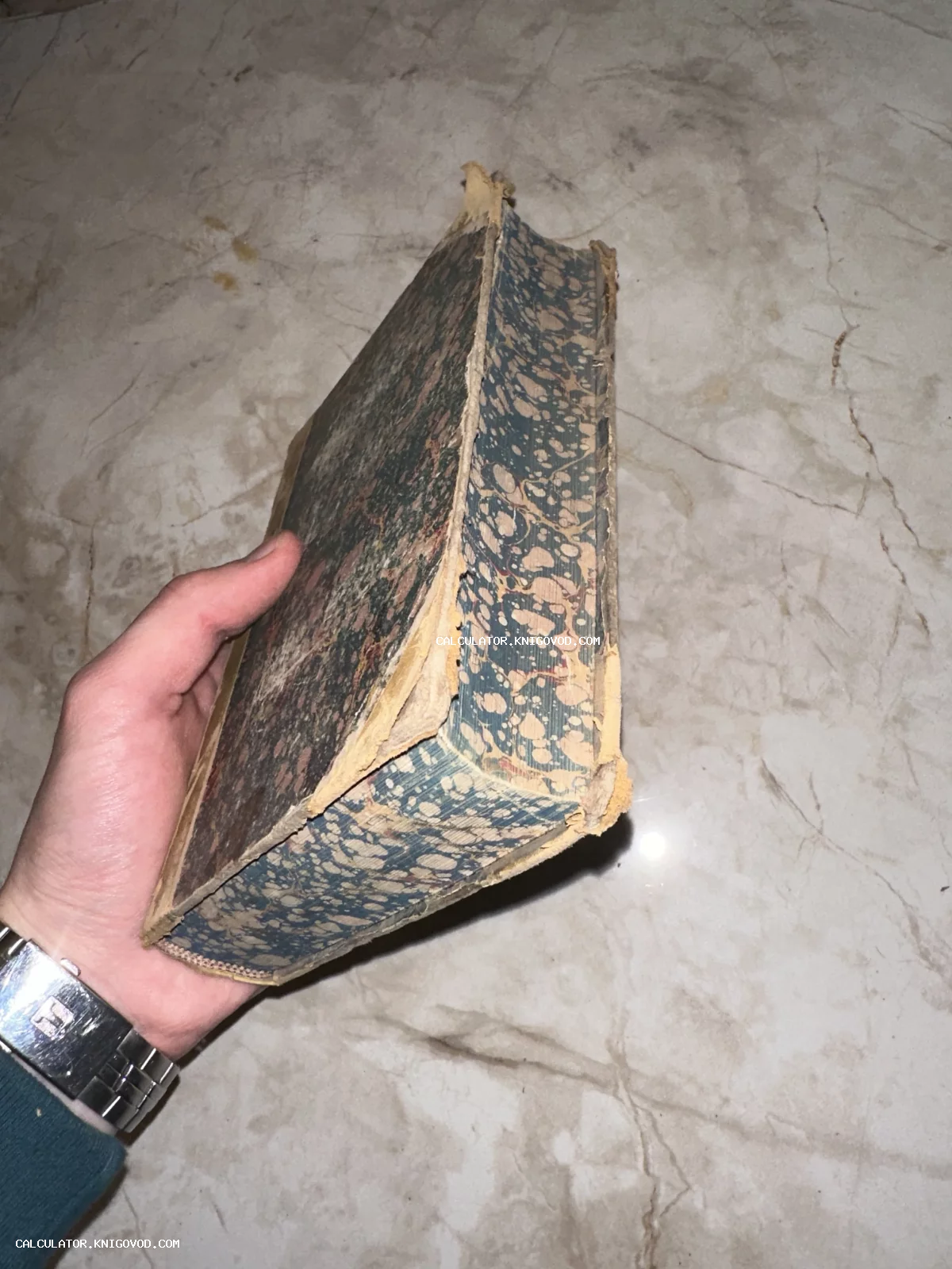 Мужская рука держит массивную старинную книгу с обложкой из сине-бежевой мраморной бумаги.
