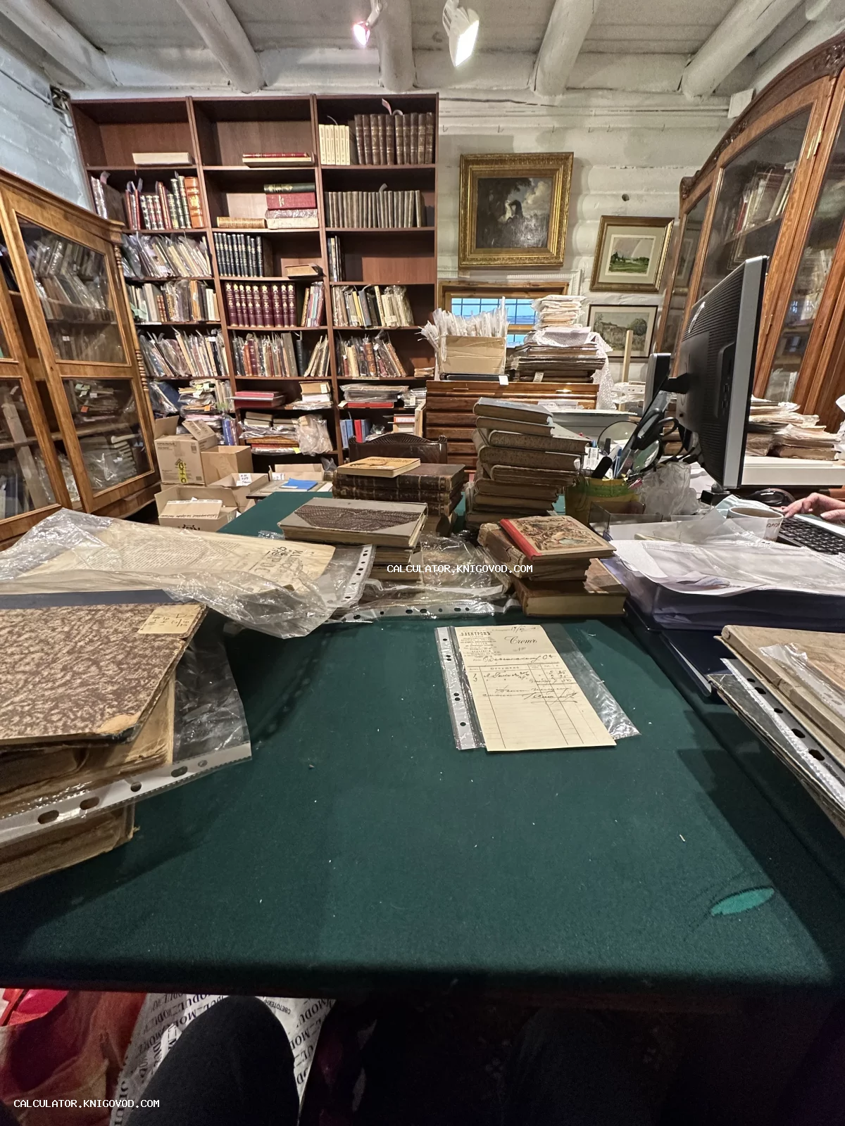 Рабочее место букиниста с множеством старинных книг на полках, стопками документов на зеленом столе и картинами в рамах на стене.