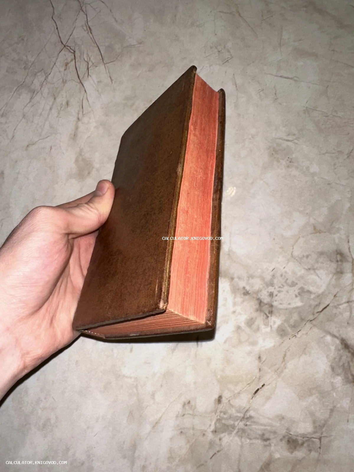 Старинная книга в коричневом кожаном переплете с красным обрезом страниц в руке на фоне мрамора