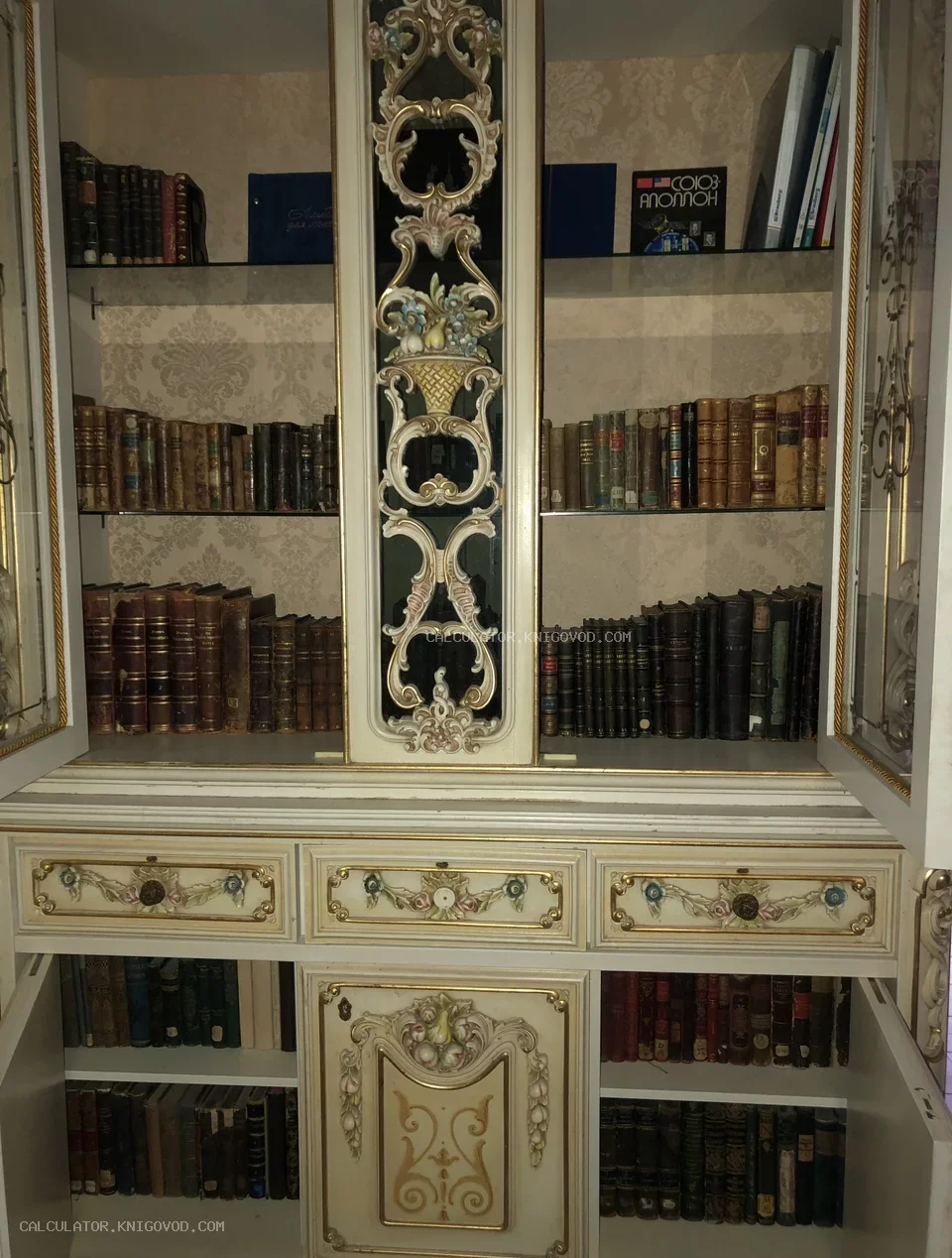 Белый антикварный книжный шкаф с золоченой резьбой и цветочным орнаментом, заполненный старинными книгами в кожаных переплетах.