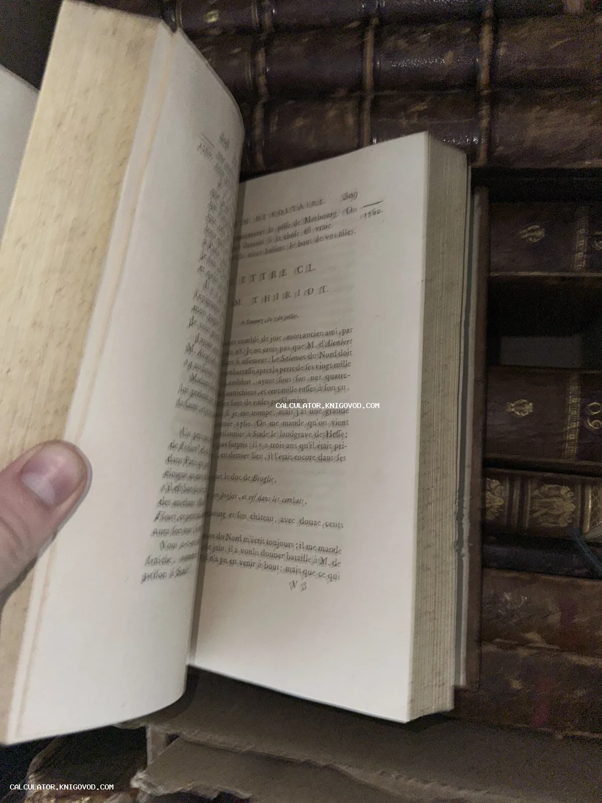 Разворот старинной книги на французском языке с текстом писем Вольтера на фоне полки с антикварными книгами в кожаных переплетах.