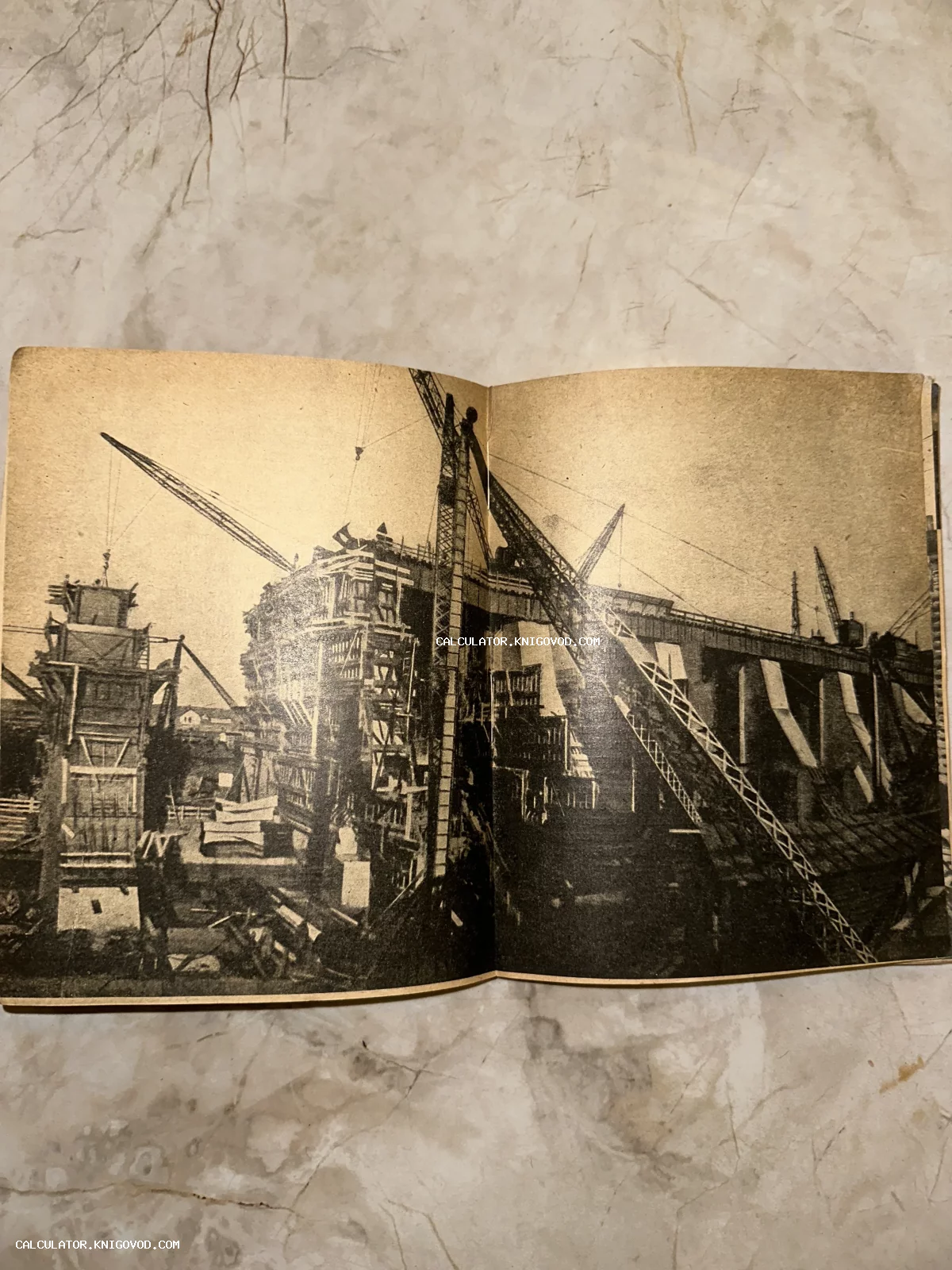Черно-белое архивное фото масштабного строительства промышленного объекта с множеством подъемных кранов на развороте старой книги.