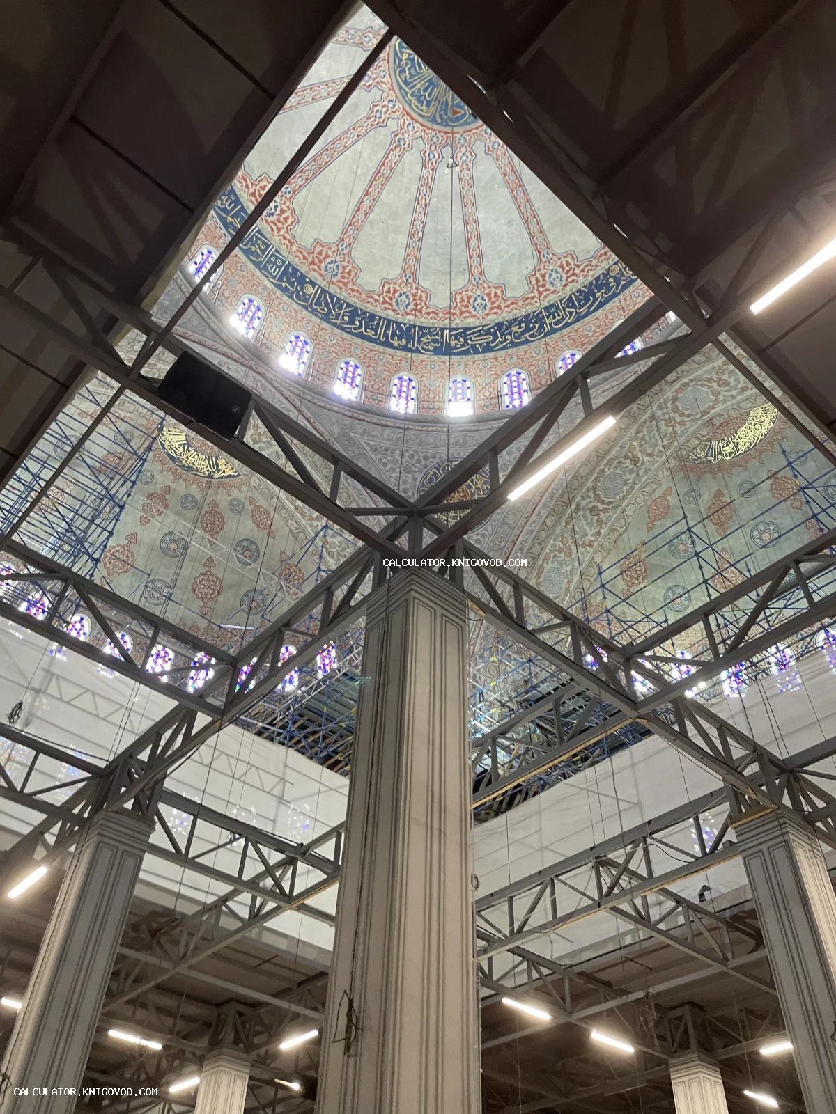 Вид снизу на расписной купол мечети с арабской каллиграфией сквозь стальные строительные леса и колонны.