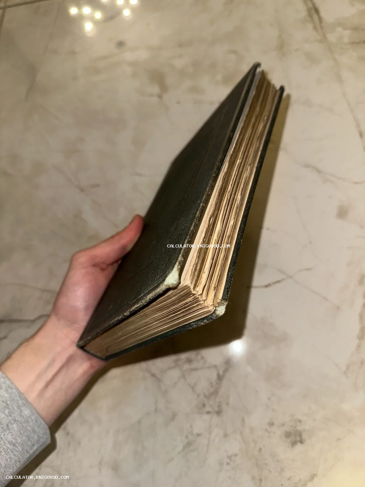 Рука держит массивную старинную книгу с пожелтевшими страницами и темной обложкой на фоне мраморного пола.
