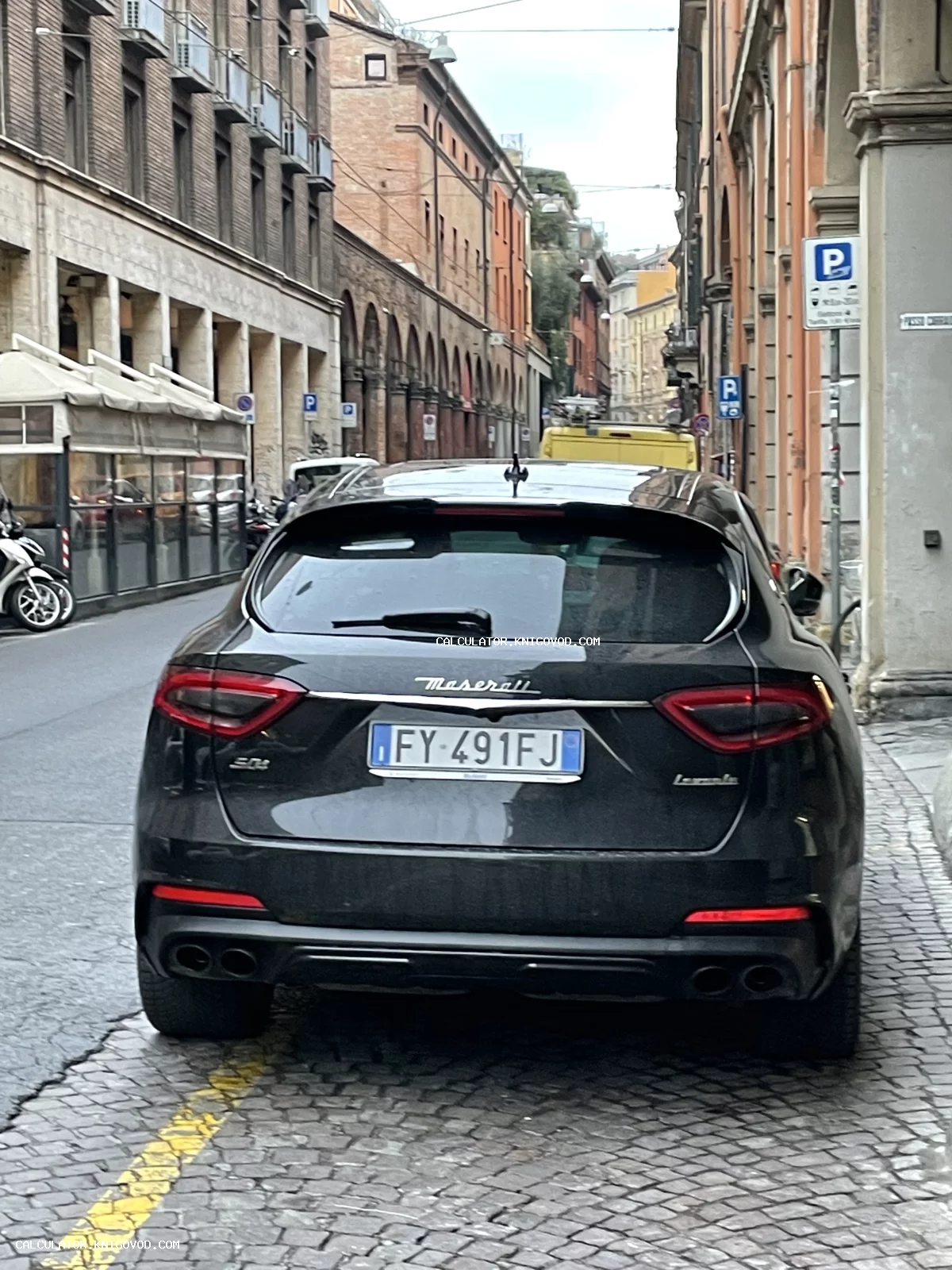 Вид сзади на черный автомобиль Maserati Levante, припаркованный на узкой мощеной улице европейского города с исторической архитектурой.