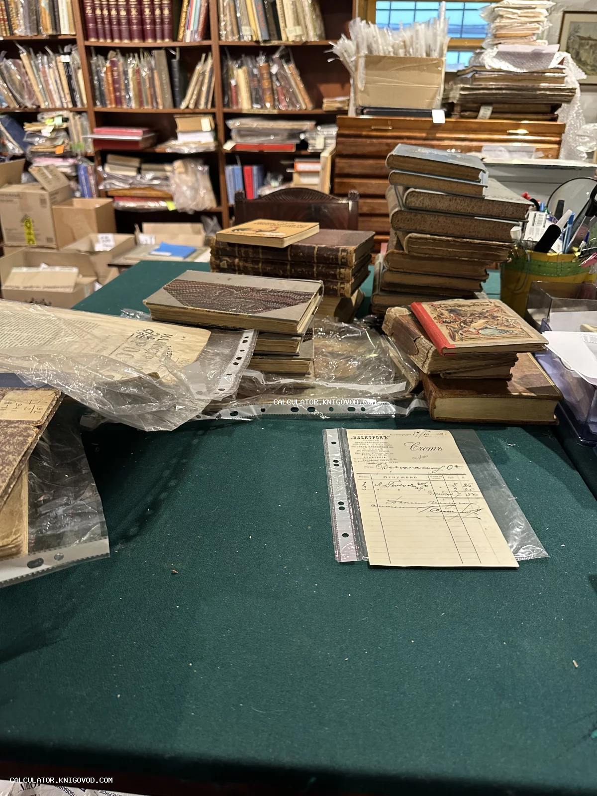 Стопки старинных книг в кожаных переплетах на зеленом столе в интерьере букинистического магазина на фоне книжных стеллажей.