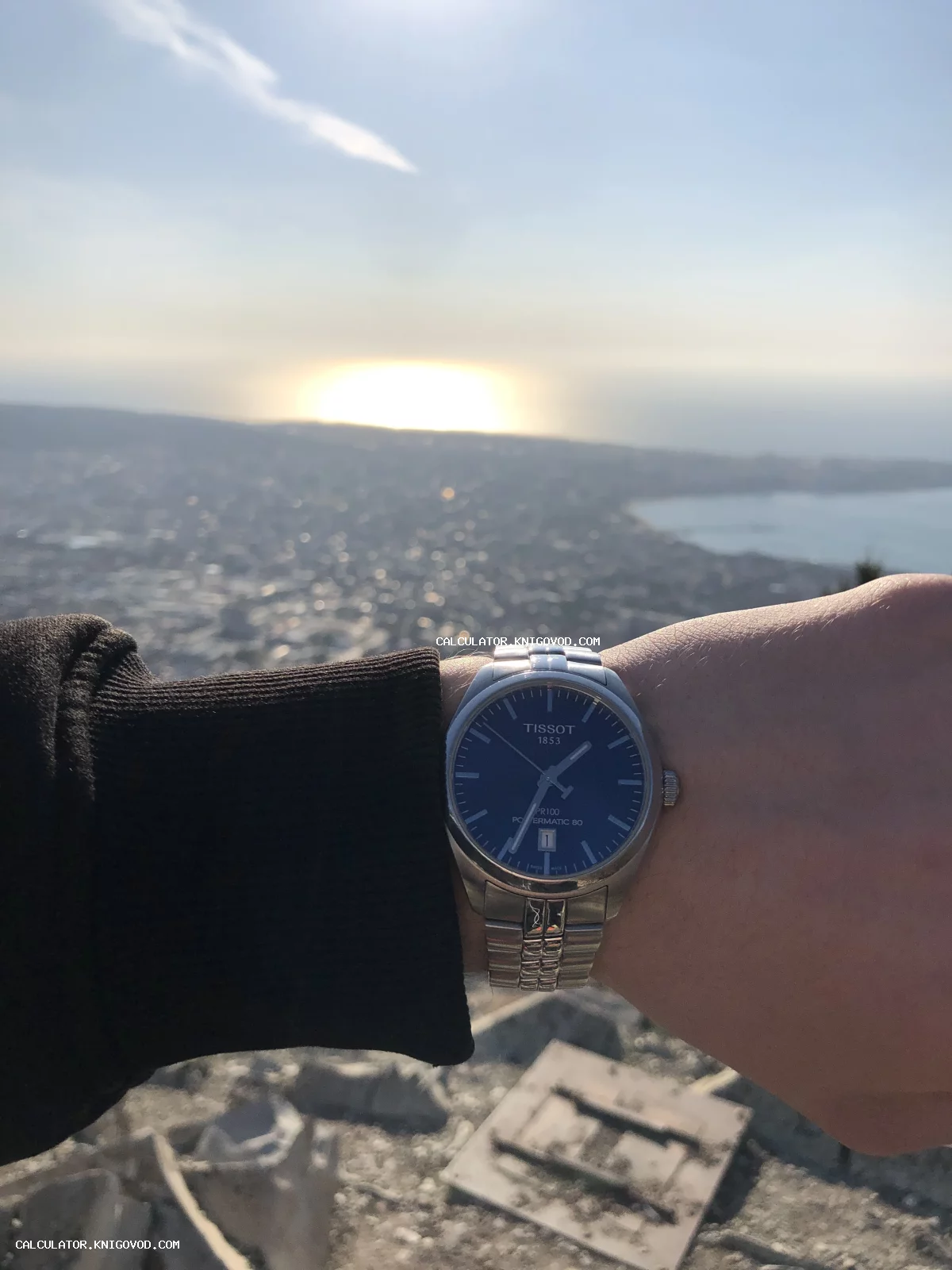 Мужские наручные часы Tissot PR100 на запястье на фоне морского побережья и города с высоты птичьего полета.