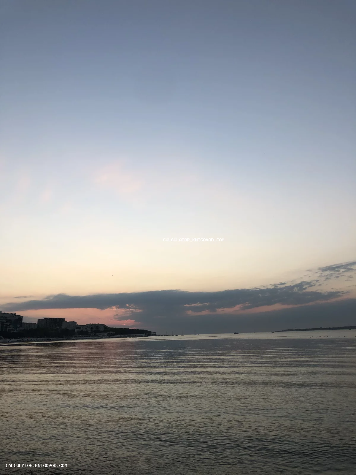 Морской пейзаж в сумерках: спокойная гладь воды, очертания городского берега и мягкий градиент вечернего неба с облаками.