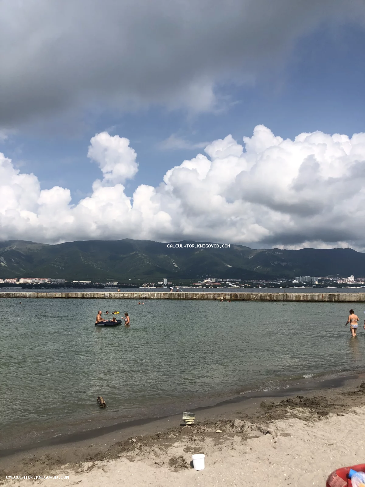 Пляж в Геленджике с видом на горы, море с купающимися людьми и волнорез под облачным небом.