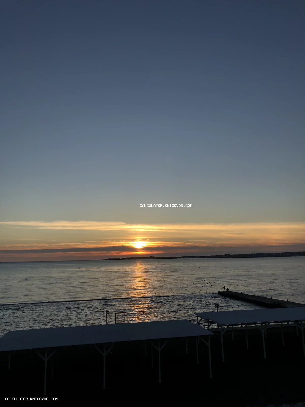 Закат солнца над морем с силуэтами прибрежных навесов и пирса в сумерках.