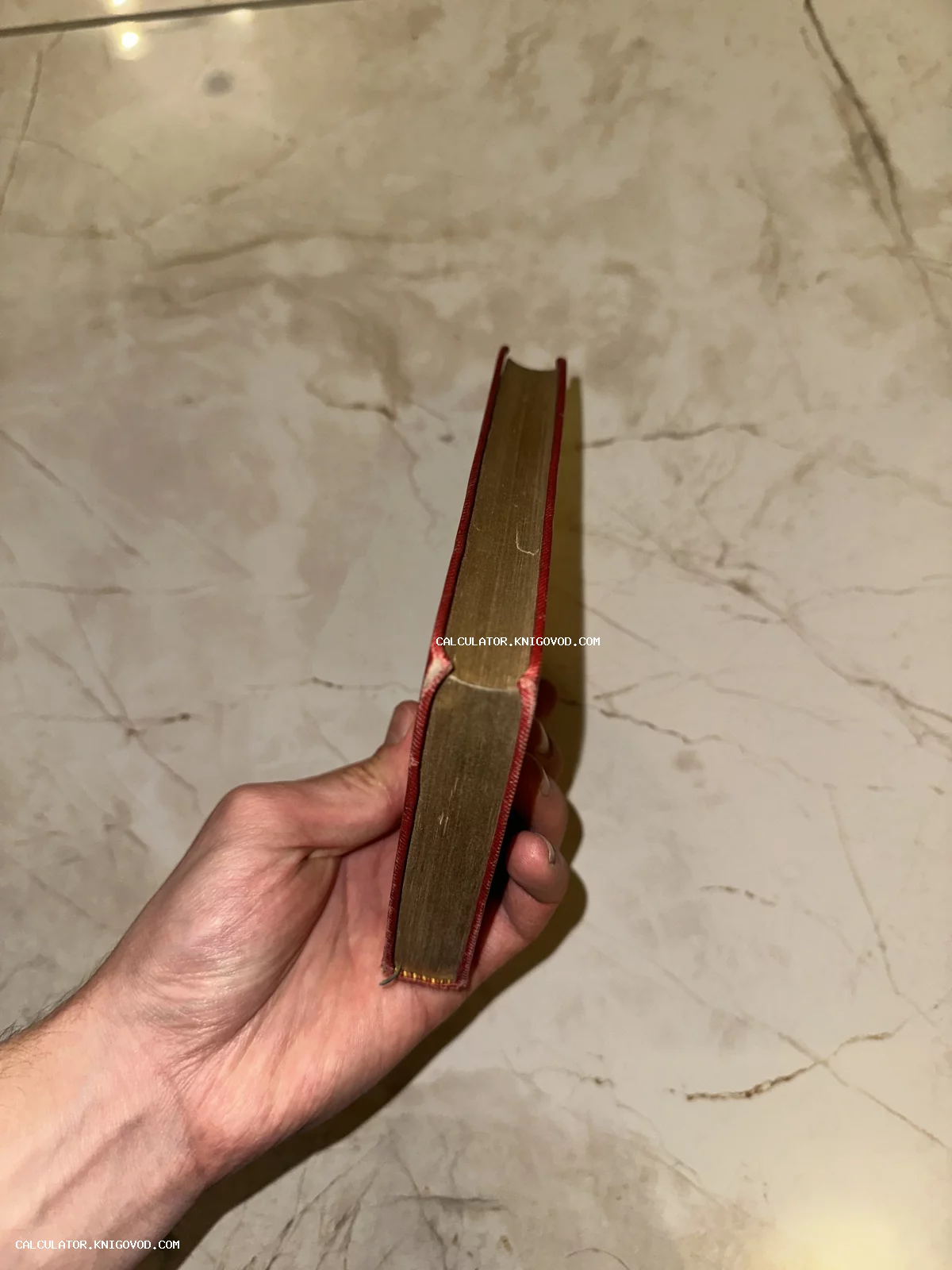 Старинная малоформатная книга в красном переплете с золотым обрезом в руке на фоне мрамора.