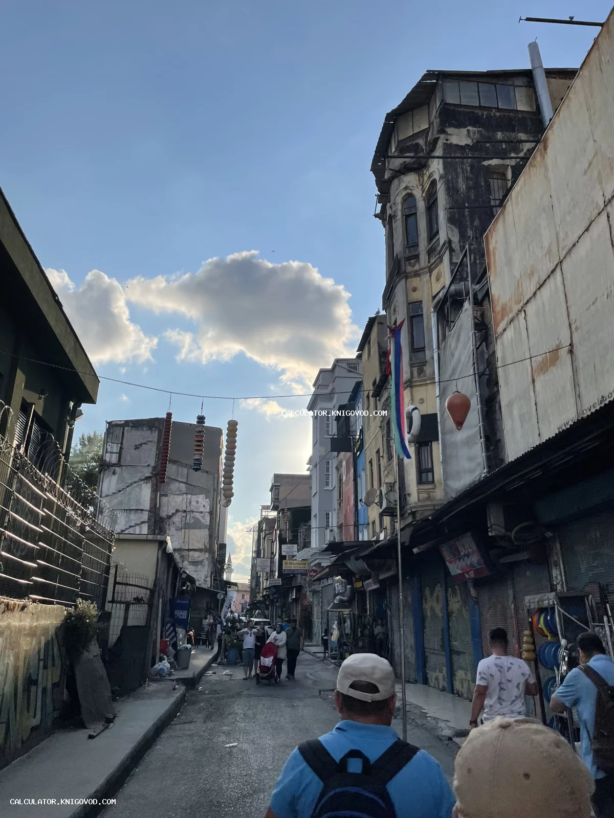 Узкая городская улица со старыми многоэтажными домами, облупившимися фасадами и прохожими под ясным небом.