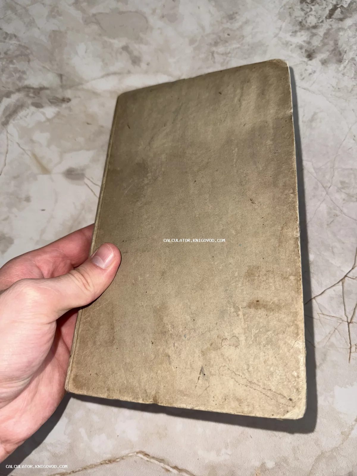 Рука человека держит старинную книгу в пожелтевшей картонной обложке без надписей на фоне мраморной поверхности.
