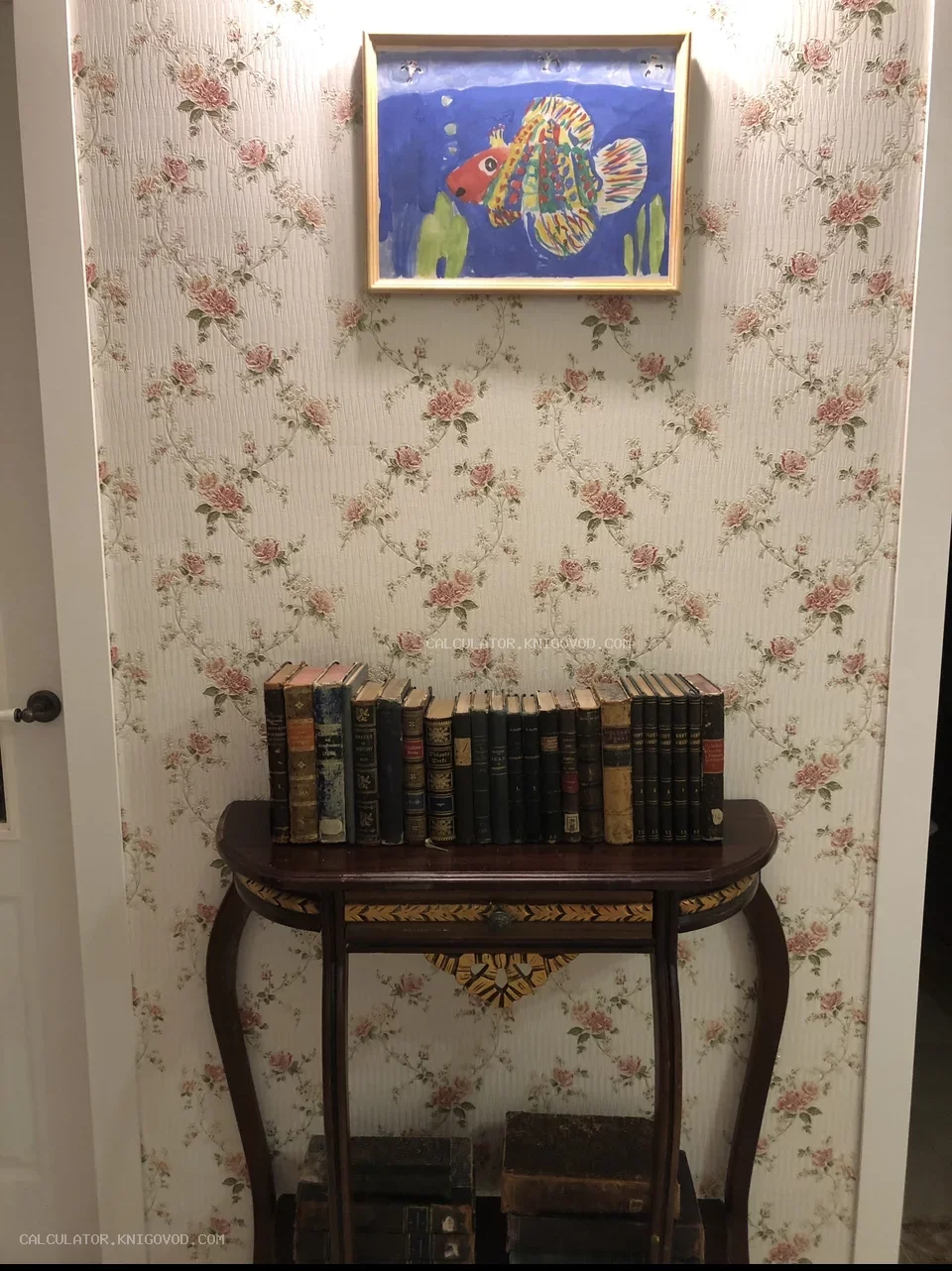 Ряд старинных книг в потертых кожаных и тканевых переплетах, выставленных на деревянной консоли перед стеной с цветочными обоями.