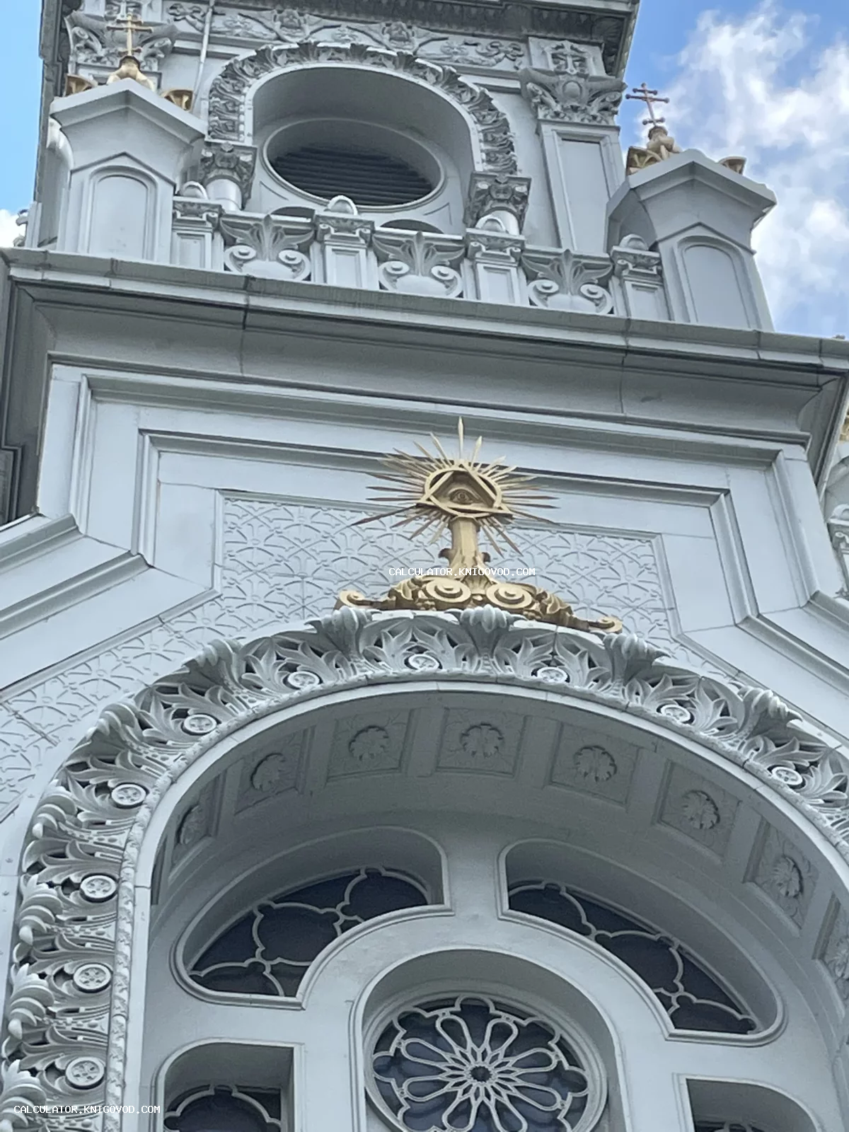 Позолоченное изображение Всевидящего ока в треугольнике с лучами на белоснежном архитектурном фасаде с арками и лепниной.