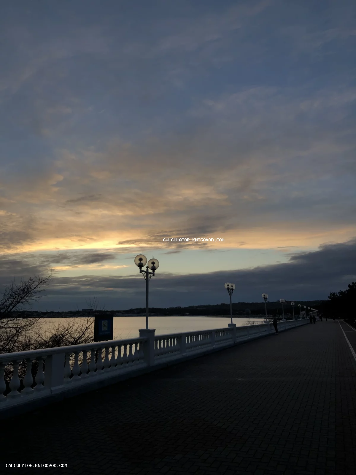 Вид на вечернюю набережную с белой классической балюстрадой, уличными фонарями и закатным небом над гладью воды.