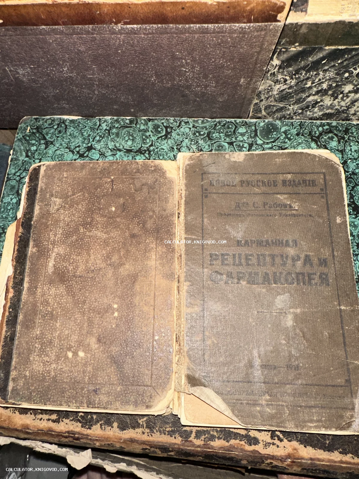 Старинная книга в потрепанном переплете с надписью Карманная рецептура и фармакопея, лежащая на мраморном фоне.